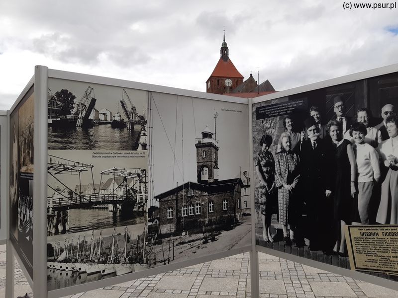 Plansze wystawy z czarno-białymi zdjęciami, nad nimi w tle wieża kościoła