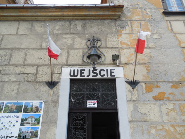 Wejście do zamku - niezbyt wielkie drzwi, nad nimi zabytkowa lampa i dwie polskie flagi